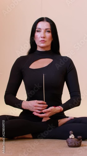 Vertical video. Meditation pose. Zen atmosphere. Woman with black hair sitting in lotus position holding burning incense stick in yoga studio.