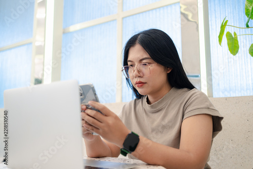 Serious Asian freelance woman wearing eyeglasses using a smartphone while sitting with a laptop in a green cafe