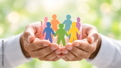 Man hands holding and protecting colorful paper cut-out people in a circle against a blurred nature background, symbolizing unity, social inclusion, diversity, community support, and teamwork.