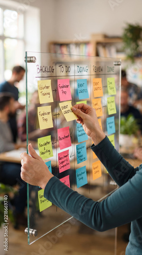 Colleague Placing Sticky Notes on Transparent Glass Kanban Board in Office