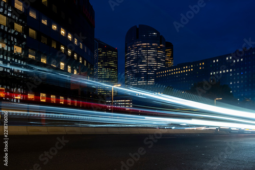 Light trails sweep through a night city beside office buildings as traffic motion crosses the street and the trails continue between the buildings and architecture
