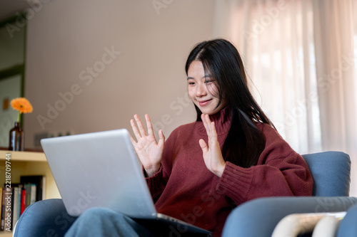 Young Asian woman waving hands, gesturing on laptop call on sofa in a living room or cafe.