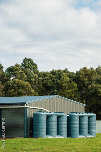 Two large slimline polyethylene rain water tanks alongside a steel shed on a farm set against a backdrop of dense eucalyptus trees under a broad, cloud-filled sky, Victoria, Australia