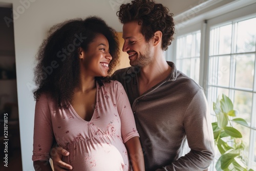 Pregnant multi ethnic couple embracing and smiling with love, standing by a bright window at home, celebrating new life, family, and the joyous anticipation of welcoming their baby