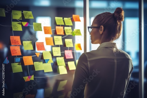 Woman standing in an office, focusing on a transparent board covered with various colorful sticky notes connected by lines, indicating a complex project management or brainstorming session