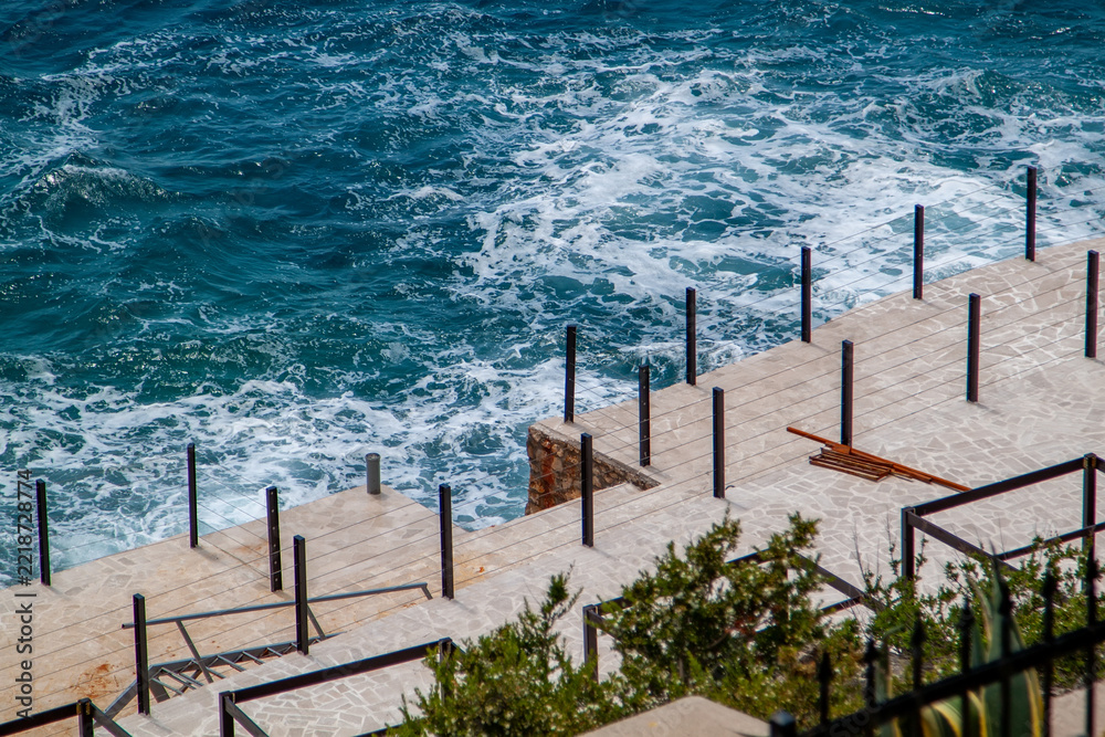Fototapeta premium High angle view of stone paved terrace overlooking deep blue sea with white waves crashing against rocky shoreline below modern metal railing fence on sunny day in Mediterranean.