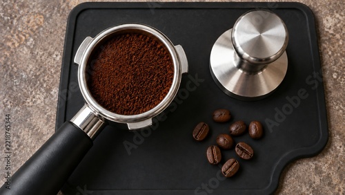 Ground coffee in a portafilter beside a steel tamper and roasted beans