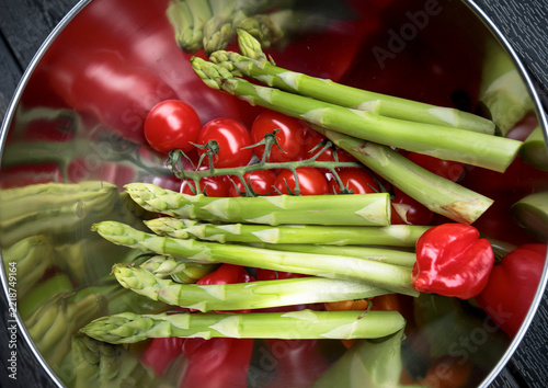 Fresh green asparagus spears with cherry tomatoes on vine in polished steel mixing bowl, vivid red green contrast, mirror reflection visible, top view food photography on dark wooden surface