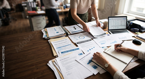 A team of office workers reviewing documents, business charts, and data reports on a busy wooden desk with a laptop