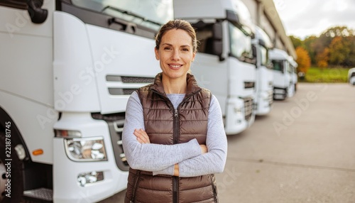 Female truck driver standing confident with crossed arms in front of fleet trucks at storage warehouse