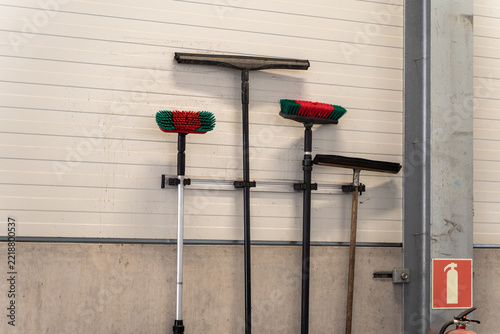 Cleaning tools including squeegees and brushes with red and green bristles stored on a wall rail in a workshop, fire extinguisher sign nearby