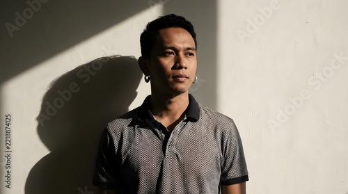 A pensive young Southeast Asian man from Indonesia wearing traditional Balinese silver hoop earrings paired with a casual patterned polo shirt, standing against a wall with dramatic sunlight shadows.