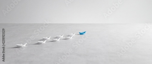 Minimalist Wide Background of One Blue Paper Boat Leading a Group of White Paper Boats on a Gray Surface, Symbolizing Leadership, Innovation, Business Vision, and Individual Success.