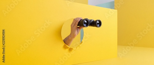 Minimalist Concept of Discovery and Research with a Hand Holding Binoculars Through a Hole in a Bright Yellow Background for Business Strategy, Future Vision, and Curiosity Marketing