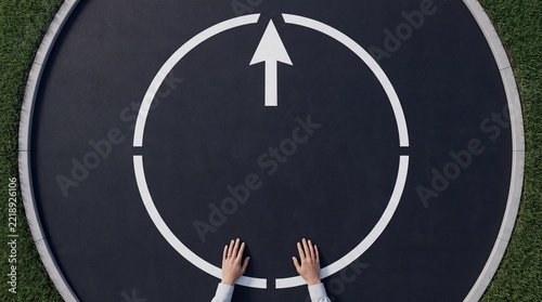 High angle overhead view of a person's hands reaching toward a circular graphic with a white arrow pointing forward on a dark textured background, symbolizing guidance and business strategy.