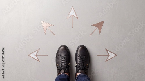 High angle view of professional person in black leather shoes standing on concrete floor facing multiple rose gold arrows pointing in different directions, symbolizing choice, decision making, and