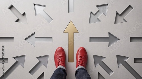 A first-person perspective of a person wearing red sneakers standing on a gray concrete floor with multiple arrows pointing in different directions and one distinct gold arrow showing the right path.