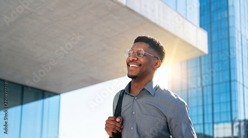 Successful Young African American Businessman with Eyeglasses Smiling and Looking Away While Walking in Front of a Modern Glass Office Building During a Bright Sunny Morning in the City
