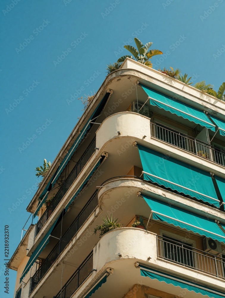 Fototapeta premium Low angle view of old apartment building with blue awnings. Mediterranean style architecture features white balconies, green plants on rooftop. Sunny day in European city with summer vibes. Urban