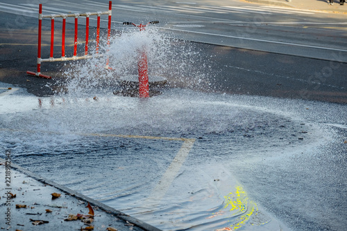 Water spraying from broken fire hydrant flooding city street, red safety barrier in background