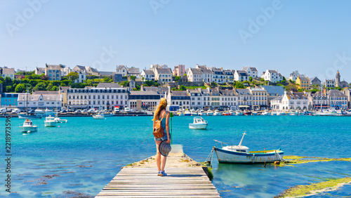 Young female traveler with backpack holding sun hat standing on wooden pier looking at coastal harbor town with boats on sunny day
