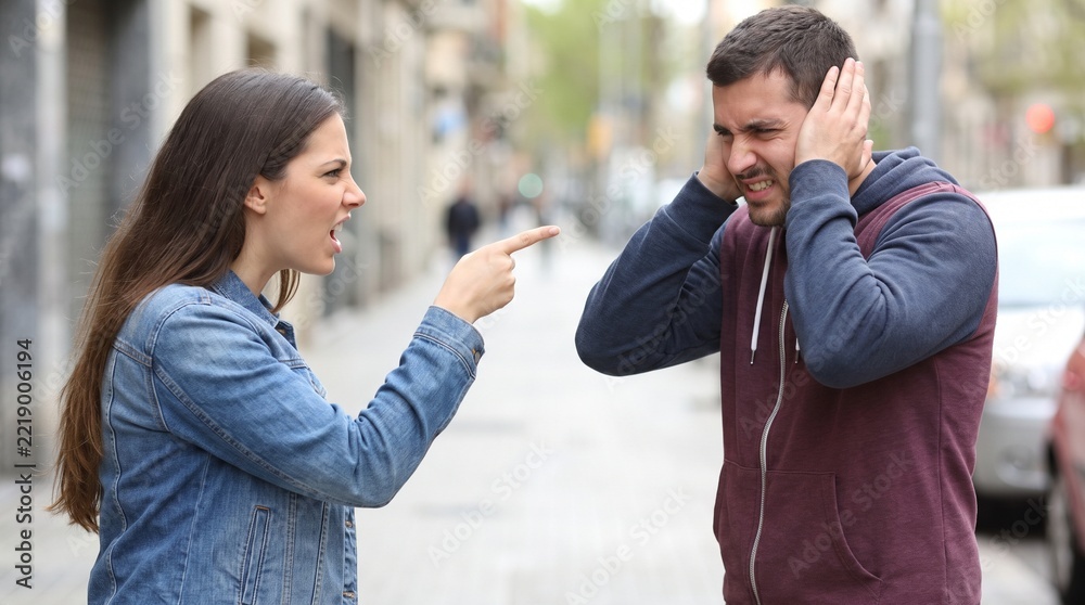 Fototapeta premium A woman angrily points her finger and yells at a man who covers his ears during a street argument.