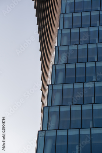 A close-up architectural photograph contrasts a modern glass façade with a projecting timber-clad structure against a pale, open sky