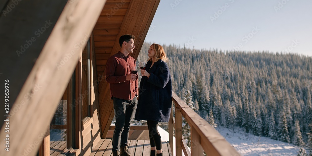 Fototapeta premium Couple enjoying wine on a snowy cabin balcony with forest view for winter vacation lifestyle banner and cozy getaway design