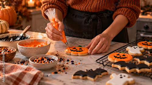 Woman decorating Halloween sugar cookies with orange icing in a cozy kitchen, Baker piping orange details on a pumpkin cookie surrounded by festive treats, Making homemade Halloween cookies 