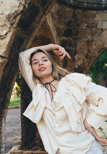 A portrait of a stylish, fair-haired woman in a beige ruffled blouse; she is posing outdoors under an old stone arch and looking straight ahead.