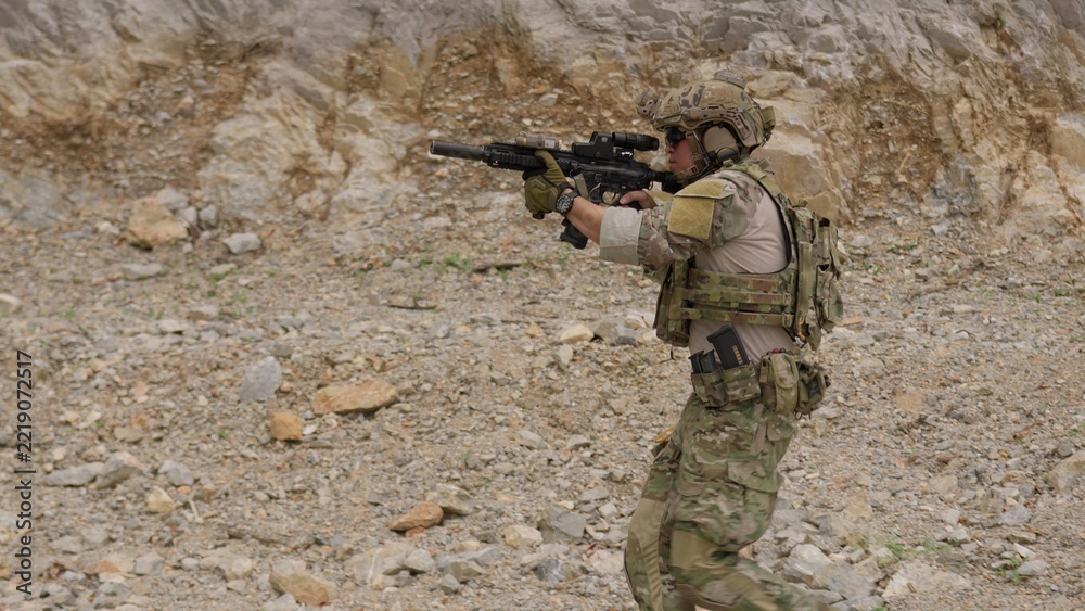 Fototapeta premium A special forces operator in full combat gear advances cautiously through a rocky, hostile environment while aiming his assault rifle on a tactical security patrol.