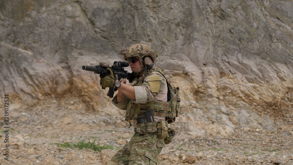Fototapeta premium A special forces operator in full combat gear advances cautiously through a rocky, hostile environment while aiming his assault rifle on a tactical security patrol.