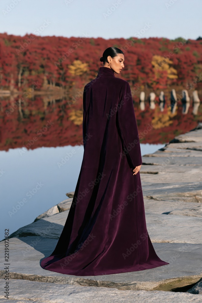 Fototapeta premium Woman wearing long purple velvet coat with flowing train walking on stone jetty over reflective lake with autumn trees for fashion editorial banner and poster