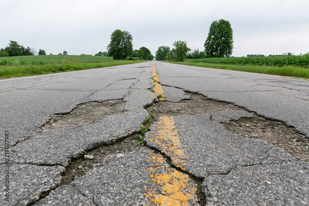 Fototapeta premium The Cracked Rural Road Stretch with Yellow Center Line and Green Fields
