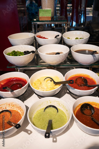 Assorted sauces, dressings, peppers, and condiments arranged in white bowls at a self-service buffet station for dining and cooking.