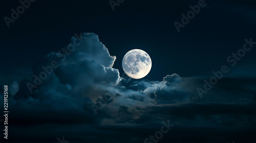 a large, full moon partially obscured by fluffy clouds. The sky is a deep navy blue, with a few scattered stars. The clouds are a mix of white and gray, with some appearing soft and fluffy.