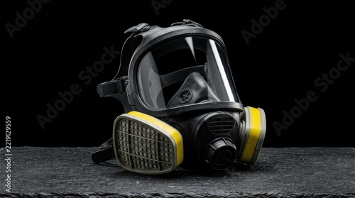 A professional black gas mask with yellow accents and clear visor rests on a dark textured surface