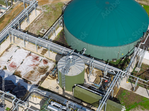 Aerial view of a biogas plant featuring a large green storage tank, a smaller cylindrical tank, and a complex network of industrial pipes on a concrete site in the United Kingdom.
