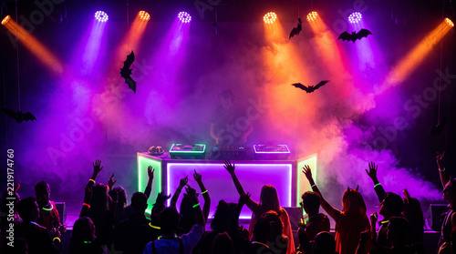 People dance at a vibrant Halloween party with purple spotlights and bat decorations.