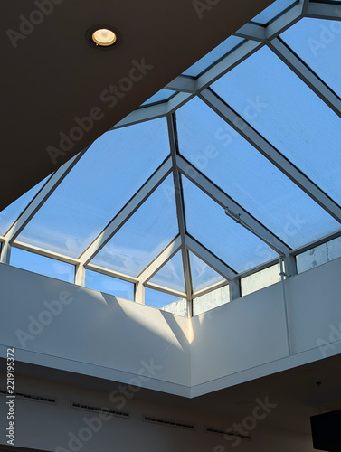 Geometric skylight structure providing natural light in a modern building interior, with a recessed ceiling spotlight and white walls.