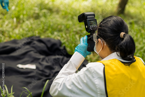 Forensic investigator in protective clothing photographs a covered object outdoors in a wooded area, documenting evidence at a field scene.