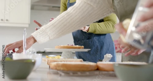 Jar being passed, aproned is scooping jam onto cake stand, partner is passing tools for stacking
