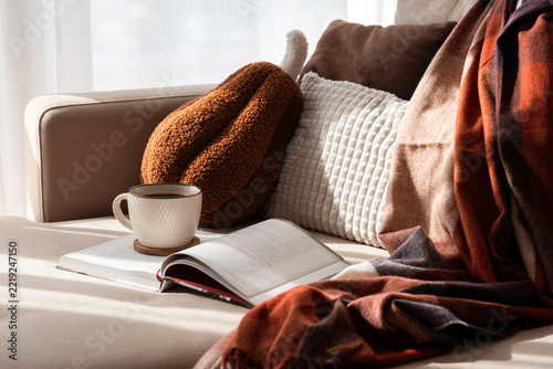 Cozy indoor scene with a warm cup of coffee and an open book on a comfortable sofa next to soft pillows and a plaid blanket.