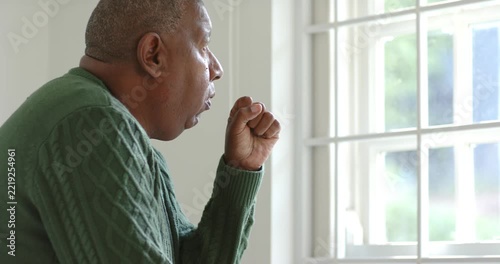 African American man sitting by window briefly coughing into fist, hand on chest calming