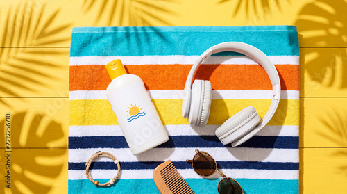 White wireless headphones and sunscreen bottle on a striped beach towel over a vibrant yellow background with soft tropical palm shadows for summer vacation concepts