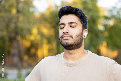 Young man with wireless earbuds meditating outdoors, eyes closed, finding calm and inner peace amid golden-hour foliage and quiet mindfulness in nature