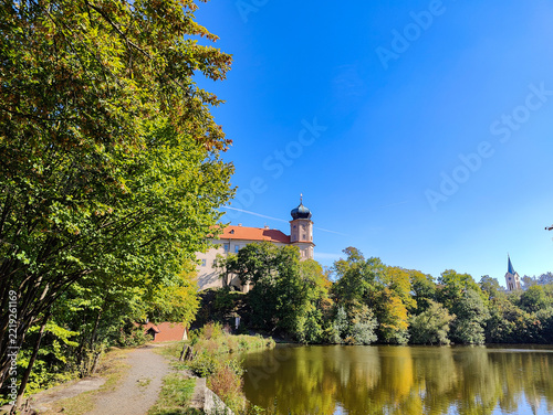 A picturesque landscape featuring historic building with red roof and dome overlooking calm lake, surrounded by vibrant green trees under bright blue sky. Gravel path winds along water's edge.