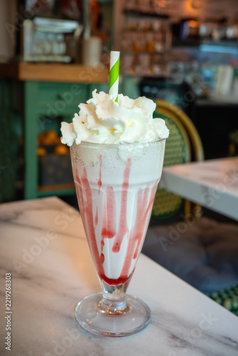 Luxury milkshake with strawberry sauce and whipped cream, in a long glass with a paper straw on a table with a marble style finish. The drink is inside a cafe.