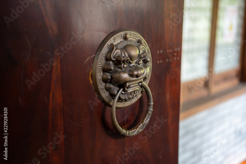 Close-up of an ornately decorated Asian door knocker in the shape of a guardian lion, mounted on a solid reddish wooden door.