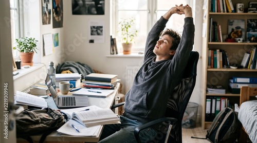 Young man stretching arms up while sitting at a messy desk in a student dorm room